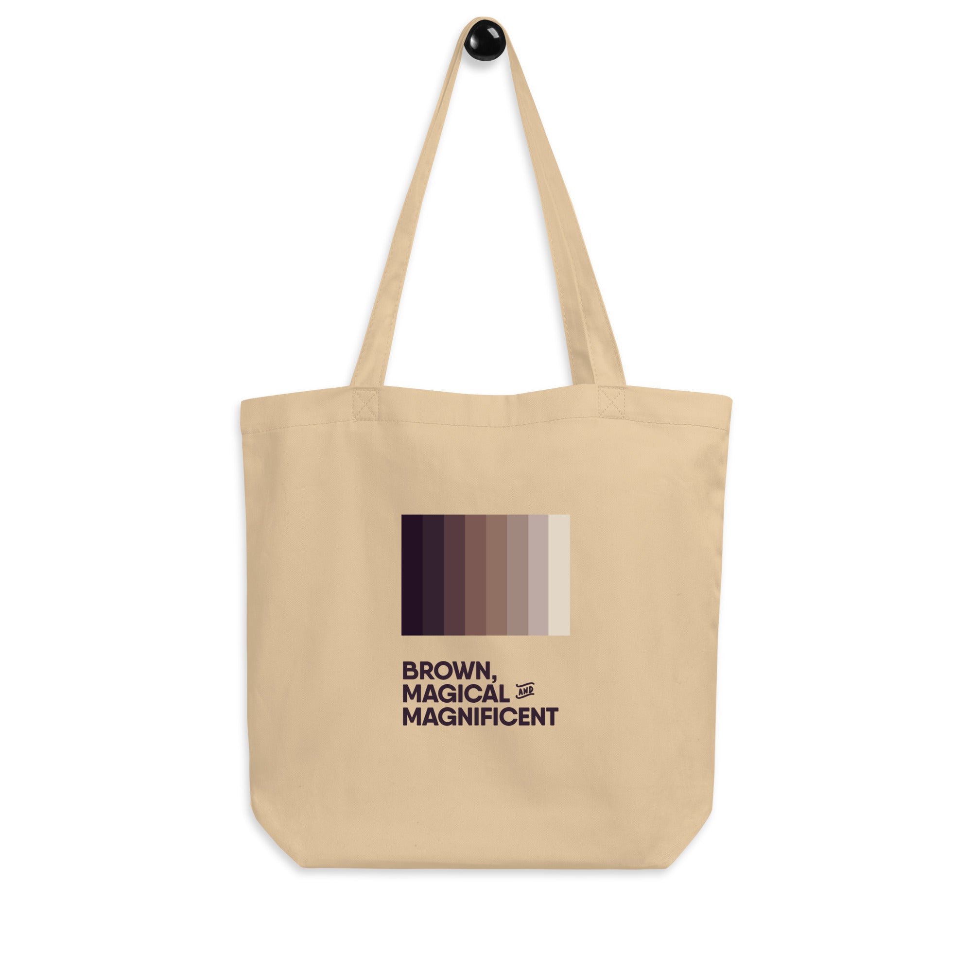 A oyster-colored canvas tote bag with sturdy handles. The words "Brown, Magical, and Magnificent" are printed in various shades of brown, creating a gradient effect.