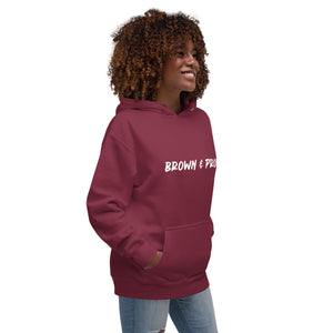 A radiant smile shines on a Black woman's face as she wears a charcoal hoodie proclaiming "Brown & Proud," capturing a moment of self-love and acceptance.