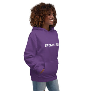 A radiant smile shines on a Black woman's face as she wears a forest green hoodie proclaiming "Brown & Proud," capturing a moment of self-love and acceptance.